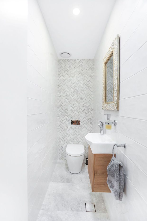 A narrow, bright contemporary bathroom with white wall tiles, pale grey floor tiles, a patterned chevron mosaic feature wall, wall-mounted timber vanity, white basin, toilet, chrome tapware, and an ornate framed mirror.