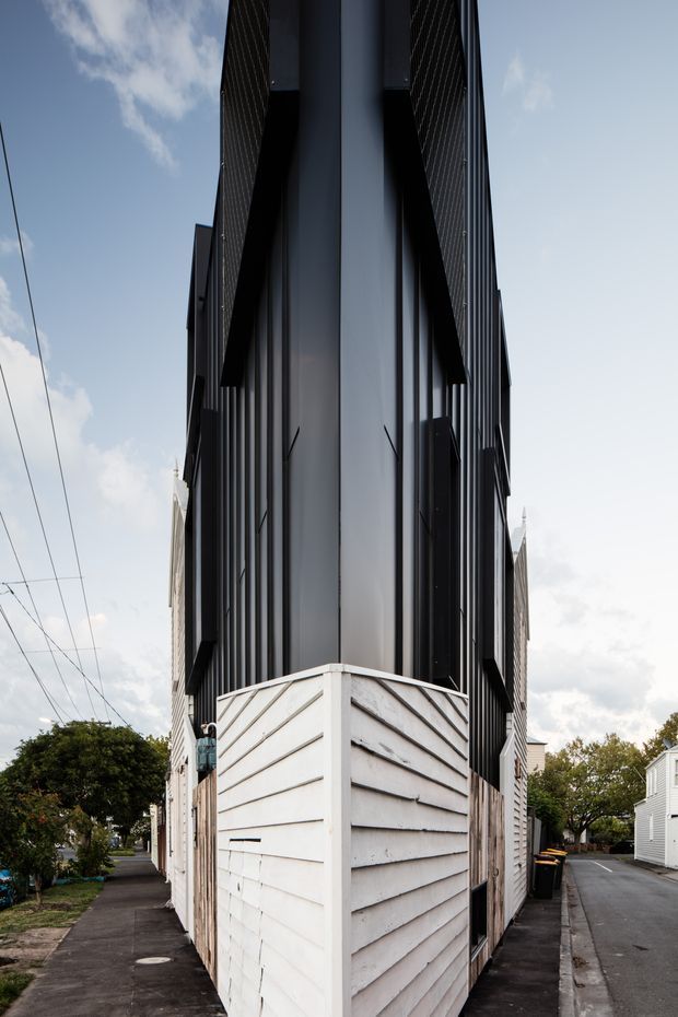 Vertical street-level view of a narrow, multi-storey urban building on a tight corner site. The facade combines dark vertical metal cladding and projecting black fins with a lower section of weathered white horizontal weatherboard cladding. Sidewalks and a narrow driveway or laneway frame the building, with neighbouring residential buildings and utility wires visible in the background.