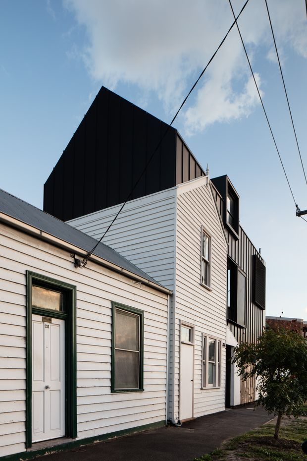 Exterior street-level view of a compact urban residential building comprising an older white weatherboard cottage in the foreground and a taller contemporary extension behind it. The extension has dark vertical cladding, a steep gabled roof form, projecting upper-level windows, and contrasting light weatherboard walls. Overhead utility wires cross the sky, with a narrow paved path and small street tree alongside the facade.