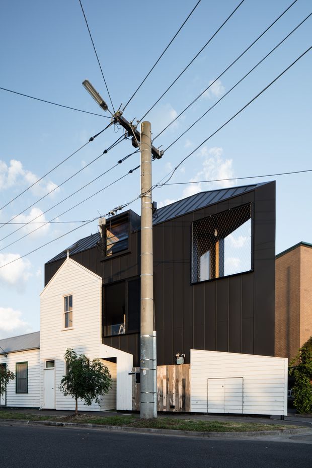 Street-facing view of a contemporary multi-storey urban residence combining a dark, vertically panelled upper facade with a white weatherboard lower extension and adjacent older white-clad house form. The building has a steep dark metal roof, large windows with mesh screening, a garage door, timber fencing, and a prominent concrete utility pole with overhead wires in the foreground.