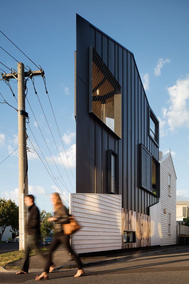 Street-level exterior photograph of a narrow multi-storey contemporary townhouse or residential building. The facade is clad predominantly in dark vertical metal panels, with a contrasting white horizontal weatherboard section, projecting windows, and a large recessed opening screened by diamond-pattern mesh. A timber-toned lower wall and white horizontal gate add contrast. Utility poles, overhead wires, pavement, and blurred pedestrians are visible in the streetscape.