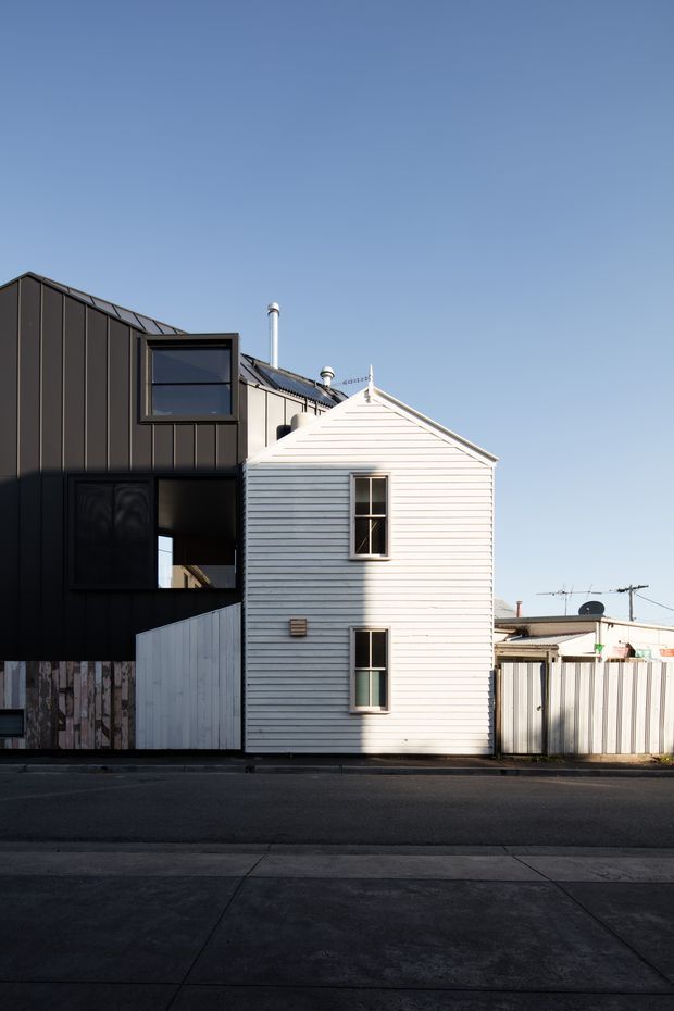 Street-facing view of a compact urban residential building, showing a dark contemporary metal-clad addition joined to a narrow white weatherboard dwelling. The composition highlights contrasting facade materials, simple gabled roof forms, vertical windows, chimneys, fencing, and the adjoining streetscape under a clear blue sky.