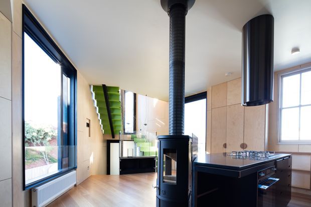 A bright contemporary open-plan kitchen and living area with light timber wall cabinetry, a dark kitchen island, large black-framed windows, and a freestanding black wood-burning stove positioned centrally in the room. A dramatic green stairway and glass balustrade are visible in the background. The space has a clean, modern fit-out with strong black, light timber, and green accents.