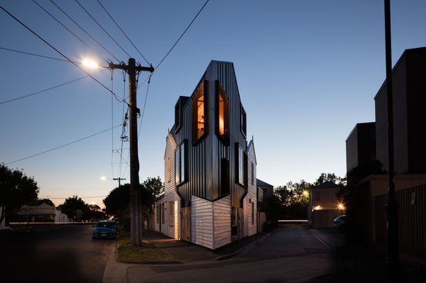 Twilight street-level view of a narrow contemporary multi-storey residential building on an urban corner site. The facade combines dark vertically ribbed metal cladding, lighter horizontal cladding and tall illuminated windows, with neighbouring buildings, driveways and overhead utility lines visible around it.