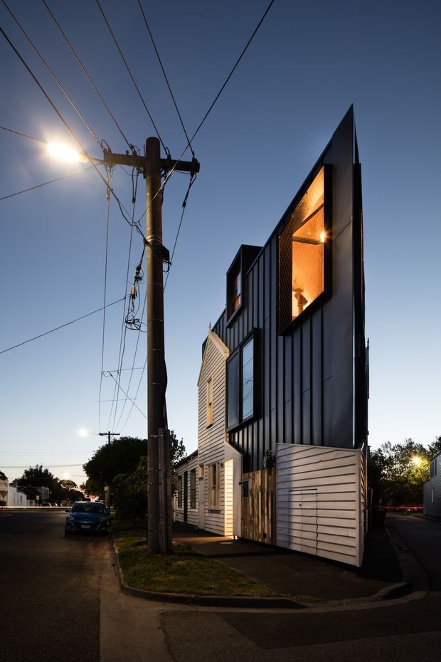 Twilight street-level view of a narrow contemporary multi-storey townhouse or infill residence. The building features dark vertically ribbed cladding, contrasting pale horizontal weatherboards, large windows, and a sharply angled projecting upper facade with warm illuminated timber-toned soffit or wall lining. A driveway or garage gate occupies the frontage, with neighbouring homes and utility poles along the residential street.
