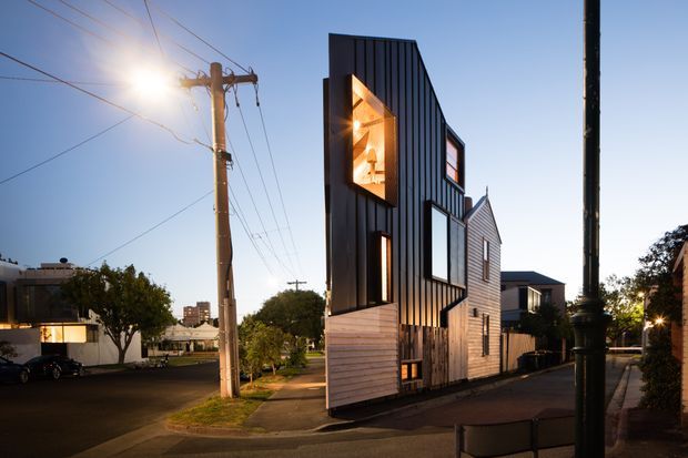 Exterior street view of a narrow, multi-storey contemporary residence on an urban corner site. The tall building has dark vertical metal cladding, pale horizontal weatherboard cladding, large windows, and a distinctive illuminated upper-level opening. Utility poles, sidewalks, neighbouring homes, and dusk lighting frame the architectural facade.