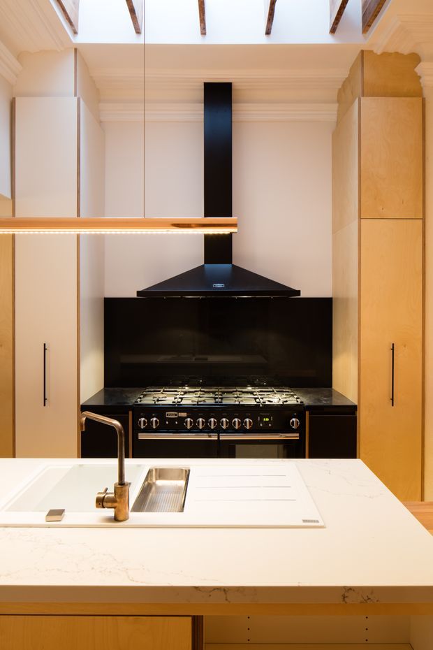 A refined kitchen focused on a large white island with integrated sink, brushed metal tap and draining board. Behind it is a professional-style gas range with black rangehood and glossy black splashback, flanked by tall light timber cabinetry. The room features warm neutral finishes, classic moulded cornices, a coffered or panelled ceiling, and exposed timber roof beams through a skylit ceiling.
