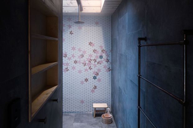 Compact, moody bathroom with a walk-in shower at the rear, a patterned floral feature wall, dark blue-grey wall finishes, a skylight, open timber shelving, and dark exposed-style towel rails. A small stool and vessel are positioned on the concrete-look floor.