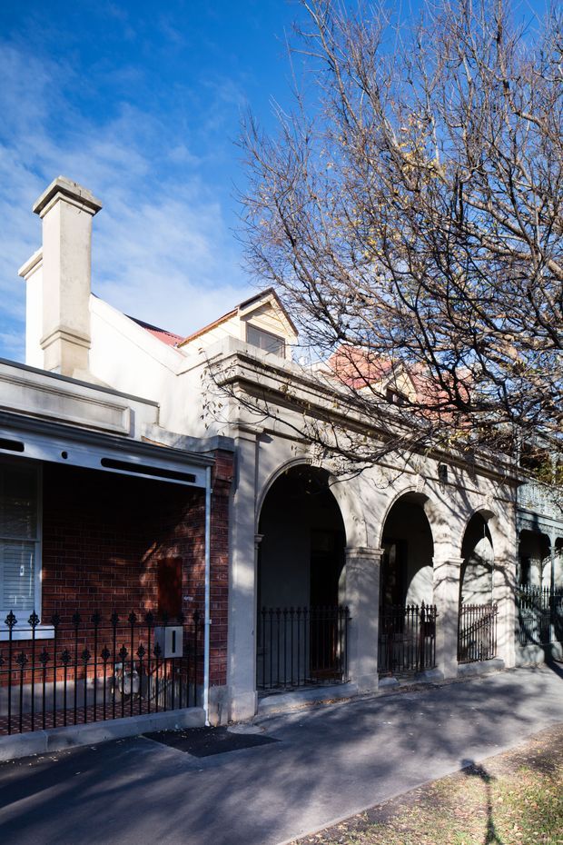 Street-facing view of a heritage-style townhouse or terrace facade with rendered masonry, exposed red-brick garage frontage, a tall chimney, red pitched roof sections, and a prominent series of arched verandah openings behind black wrought-iron fencing. Leafless tree branches cast shadows across the facade and pavement in bright daylight.