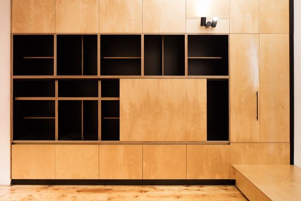 A built-in timber wall storage and media unit with warm plywood cabinetry, open black shelving, enclosed cupboards, and a projecting low platform or bench at the right. The joinery spans floor to ceiling and sits above a light timber floor, creating a clean, highly integrated interior feature.