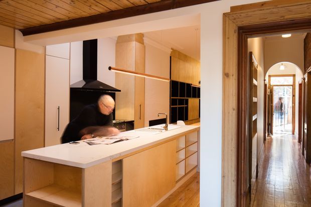 Warmly lit open-plan kitchen viewed from an adjacent interior space, with a large pale benchtop island, light timber cabinetry, black rangehood and cooktop, timber ceiling and flooring, and a rustic timber-framed doorway leading to a long arched hallway.