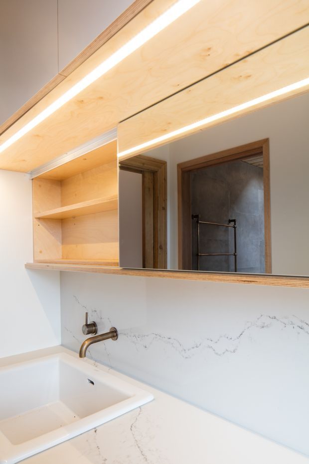 Close-up view of a contemporary bathroom vanity with a white integrated or undermount basin, wall-mounted aged-bronze tapware, pale engineered-stone vanity surface and matching marble-look splashback. A large mirrored cabinet with open pale plywood shelving and concealed linear lighting spans the wall. Timber-framed doorways and a reflected towel rail are visible in the mirror.