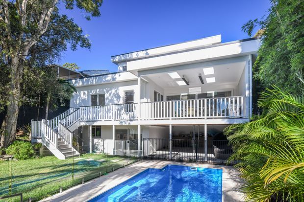 Bright contemporary two-storey residential facade with a broad covered upper balcony, white vertical balustrades, open stairway, flat roof forms and extensive glazing. A vivid blue swimming pool occupies the foreground, with landscaped planting surrounding the house.