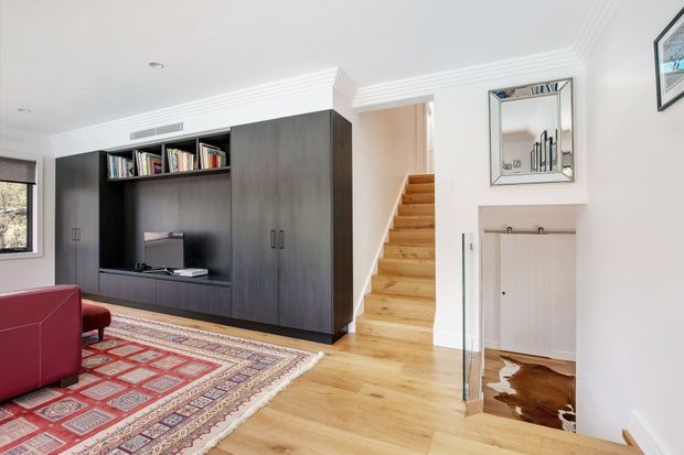 Bright, refined living room with pale timber flooring, a large dark built-in media and bookcase wall, a red upholstered sofa, and a patterned area rug. A timber staircase rises beside the joinery, while a framed mirror and concealed storage niche add detail to the white-trimmed interior. Decorative ceiling mouldings and recessed lighting contribute to a polished transitional character.