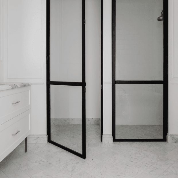 Bright bathroom interior with white paneled walls, pale marble-patterned flooring and skirting, a white vanity with slim metal handles, and black-framed glass shower screens and door. The shower enclosure includes white subway wall tiles and a visible showerhead, creating a refined monochrome look.