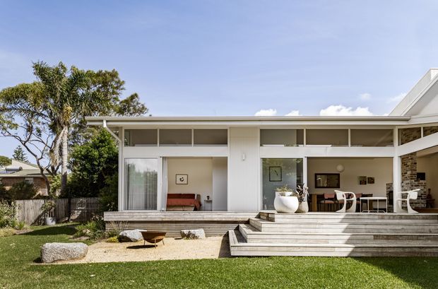 Single-storey contemporary residential home viewed from the landscaped front or rear garden. The white rendered facade has extensive floor-to-ceiling glazing, high clerestory-style windows, a low-pitched roof and a broad timber deck with wide steps. Open-plan living and dining spaces are visible through the glazing, with a brick fireplace wall at the right. The setting includes a lawn, gravel landscaping, mature trees and a fenced suburban garden.