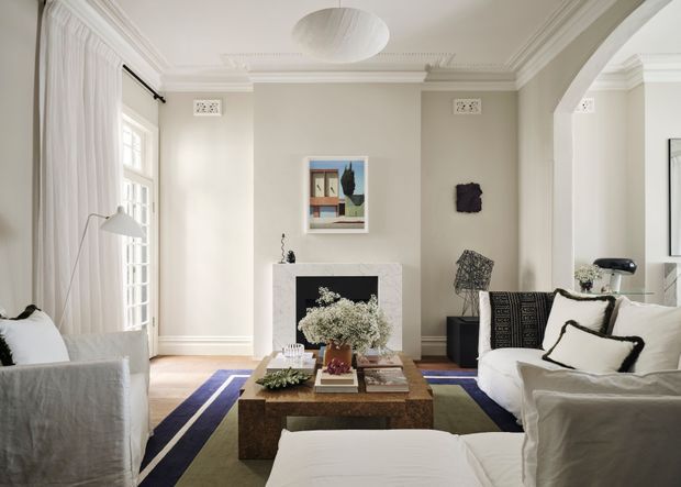 Bright, elegant living room with white slipcovered sofas and armchairs arranged around a substantial timber coffee table, a geometric area rug, and a central fireplace with a pale stone or rendered surround. The room features tall curtained windows, ornate ceiling cornices and wall vents, a large arched opening, pale neutral walls, layered artwork, and restrained contemporary furnishings within a traditional architectural shell.