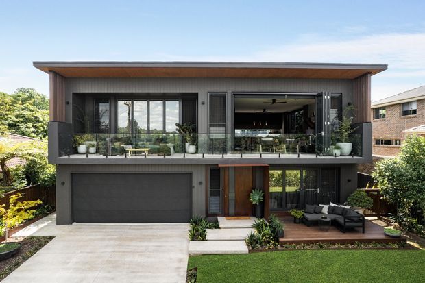 A contemporary two-storey suburban residence with a strong horizontal form, dark cladding, warm timber soffits, extensive upper-level glazing and a recessed balcony. The lower level includes a large integrated garage, timber entry door and landscaped front garden with an outdoor seating area. A partially open upper-level living space is visible behind the glazing.