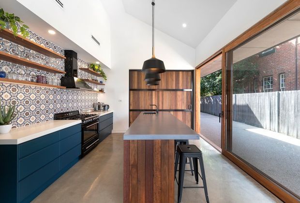 Bright contemporary kitchen with a central timber-clad island, dark blue base cabinetry, patterned ceramic tile splashback, open timber shelving, and a black statement pendant light. Full-height timber-framed glazing opens views to an enclosed outdoor area, while polished concrete flooring and warm wood finishes create a refined industrial character.