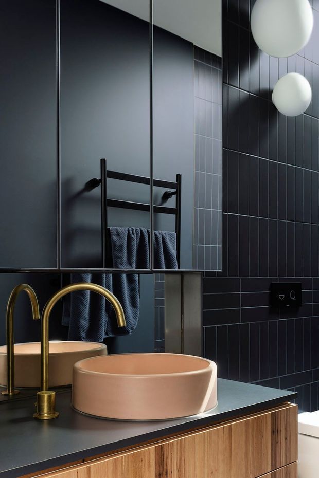 A moody contemporary bathroom featuring a freestanding round blush-pink vessel basin on a dark benchtop, brushed brass gooseneck tapware, and warm timber vanity cabinetry. Large mirrored panels reflect a black towel ladder with dark towels. The walls are clad in dark charcoal vertical and horizontal tile, with globe wall lights and black flush controls creating a refined, graphic interior.