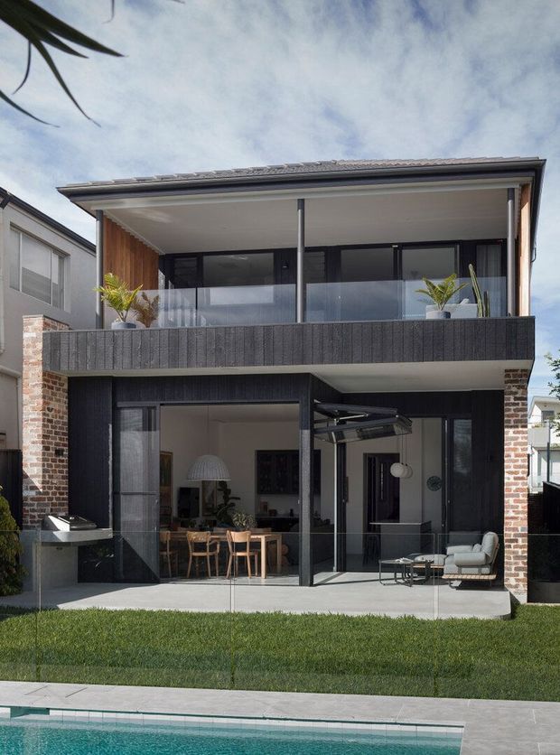 A contemporary multi-storey townhouse exterior with a dark charcoal facade, exposed red-brick side walls, broad glazed openings, and a covered upper balcony with glass balustrade. The ground level opens to a furnished outdoor entertaining area and dining space, with a neatly landscaped lawn and swimming pool in the foreground.