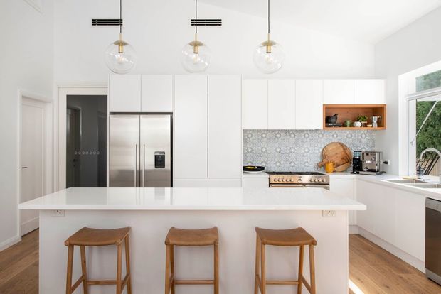 Bright contemporary kitchen with a large white island and three timber bar stools, white floor-to-ceiling cabinetry, stainless-steel refrigerator and range, patterned tiled splashback, open timber display niche, pendant globe lights, and warm timber flooring. A window and partial sink area are visible on the right.