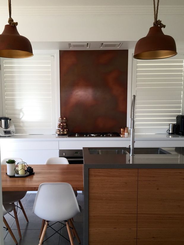 A bright, compact open-plan kitchen and dining area. The kitchen features white cabinetry, white plantation shutters, a stainless-steel sink and appliances, a timber island with dark benchtop, and a copper-toned splashback. Two large rust-coloured pendant lights hang above the island, while a timber dining table with white moulded chairs occupies the foreground.