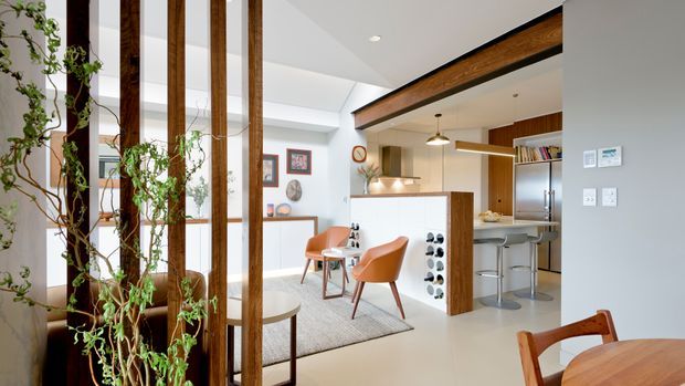 Bright open-plan residential interior showing a contemporary kitchen and dining area. A timber slat divider with trailing greenery separates the foreground from the room, while the kitchen features a white island with timber detailing, integrated appliances, pendant lighting, timber cabinetry, and bar stools. A dining table, upholstered chairs, artwork, and shelving are visible in the adjoining space. The room has a light neutral palette with warm timber accents and a partially exposed timber beam.