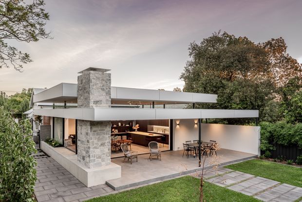 Exterior view of a contemporary multi-level residence with strong horizontal rooflines, broad open glazing, and a prominent stone-clad chimney or structural tower. The home opens onto a paved terrace with outdoor dining and an outdoor kitchen or bar visible beneath the cantilevered upper level. A landscaped lawn, stepping-stone path, mature trees, and boundary planting surround the house.