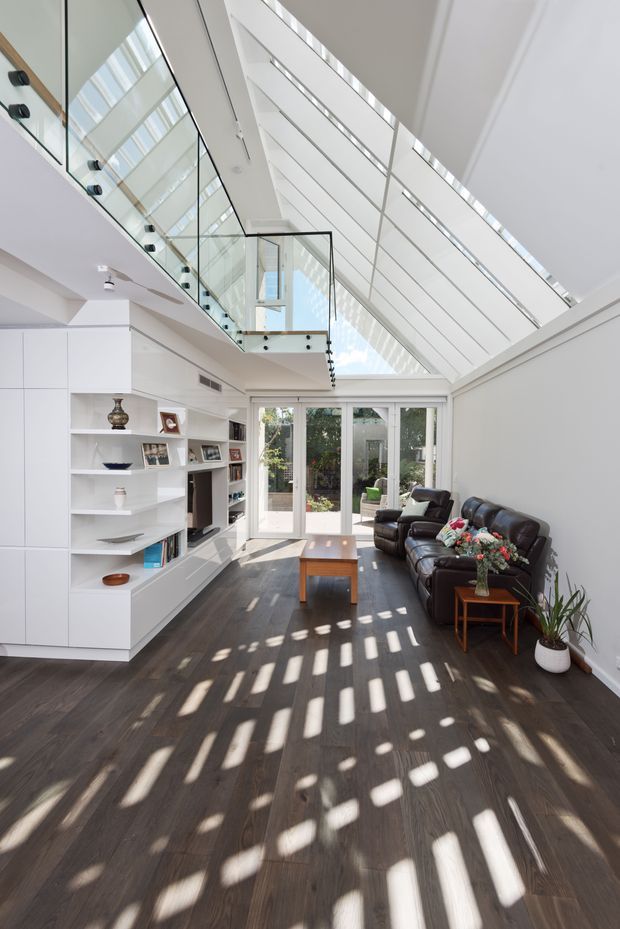 Bright contemporary living room with a dramatic glazed pitched roof and clerestory-like skylight structure. The room features dark timber flooring, white built-in cabinetry and open shelving, a large black leather sectional sofa, a timber coffee table, and full-height glazed doors opening to a garden. A glazed stair or upper-level balustrade overlooks the space, while strong patterned sunlight falls across the floor.