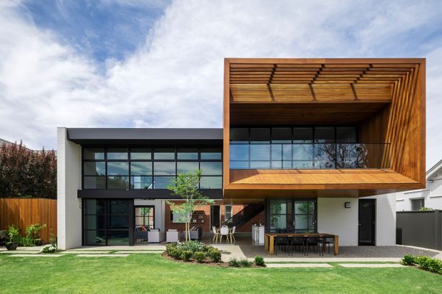 A contemporary two-storey residential facade with a bold projecting timber-clad upper-level box and deep framed terrace or window opening. The home combines dark aluminium-framed glazing, white rendered walls, timber screening and cladding, and a landscaped front lawn with a paved entry path and outdoor dining area. The projecting timber frame incorporates horizontal slatted shading beneath its soffit, creating a strong geometric architectural feature.