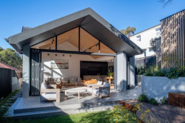 A contemporary single-storey pavilion-style extension opening onto a landscaped outdoor area. The structure has a broad pitched roof with deep eaves, exposed timber soffit panels, extensive glazed openings, dark metal framing, and an open-plan living area furnished with sofas and low tables. Concrete paving, raised planter beds, timber bench seating, and garden planting surround the space.