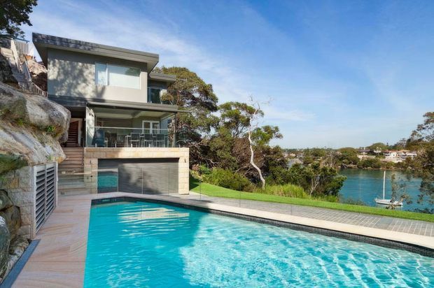 Exterior view of a modern multi-level waterfront residence built into a rocky, sloping site. The home has flat roof planes, extensive glazing, concrete-toned facade elements, a covered terrace, and a large swimming pool in the foreground overlooking a calm inlet.