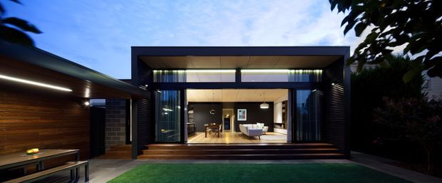 Twilight exterior view of a sleek contemporary single-storey home extension or pavilion opening onto a landscaped lawn. The dark rectilinear facade has extensive floor-to-ceiling glazing, deep recessed openings, horizontal timber-clad wall sections, and broad timber steps leading into a warmly lit living area. A covered side structure with linear lighting and timber detailing extends from the main volume.
