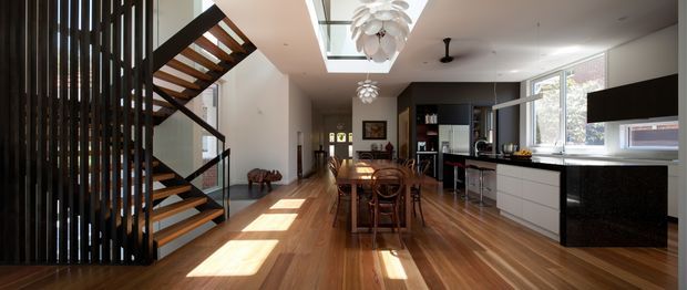 Open-plan contemporary interior combining a kitchen, dining area and stairway. Warm timber flooring and open-riser timber stairs contrast with dark vertical screening, black cabinetry and white walls. A long dining table sits centrally beneath decorative pendant lights, while the kitchen features a dark island, white cabinetry and large windows. Natural light creates strong rectangular highlights across the floor.