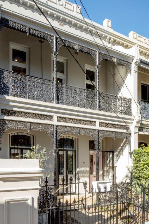 Sunlit close-up of a historic multi-storey terrace-house facade with cream rendered walls, ornate cast-iron verandah posts, lacework brackets, decorative balcony balustrades, arched ground-floor openings, and a small front garden enclosed by iron fencing. Overhead utility wires cross the view.