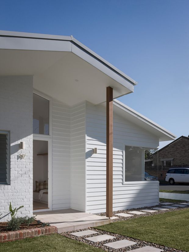 Exterior view of a compact, single-storey contemporary house with crisp white horizontal weatherboard cladding, a low-pitched skillion-style roof, white-painted brickwork, and a covered entry supported by a timber post. A paved stepping-stone path crosses the lawn to the porch, with neighbouring homes visible in the background.