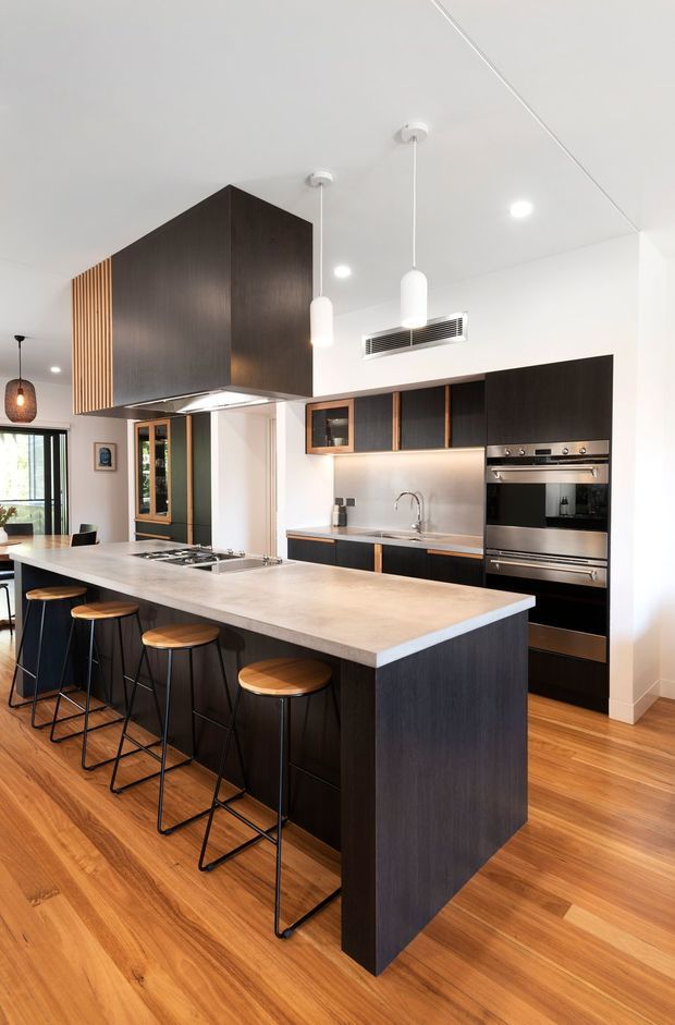 A bright contemporary kitchen featuring a large island with a pale stone benchtop and dark timber cabinetry, integrated stainless-steel appliances, a stainless splashback, and timber flooring. The island includes seating for several people on black-framed stools with timber seats. White pendant lights and recessed ceiling lights illuminate the space, while timber slatted detailing adds visual interest near the rangehood and adjoining joinery.