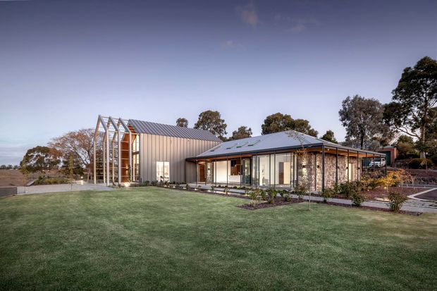 A contemporary single-storey rural residence viewed from the landscaped front lawn at dusk. The home combines a low-pitched metal roof, dark-framed glazing, vertical metal cladding, and a prominent glazed gable-like entry volume with exposed framing. A long glazed living wing opens toward the lawn, with paved paths and planted garden beds around the house.