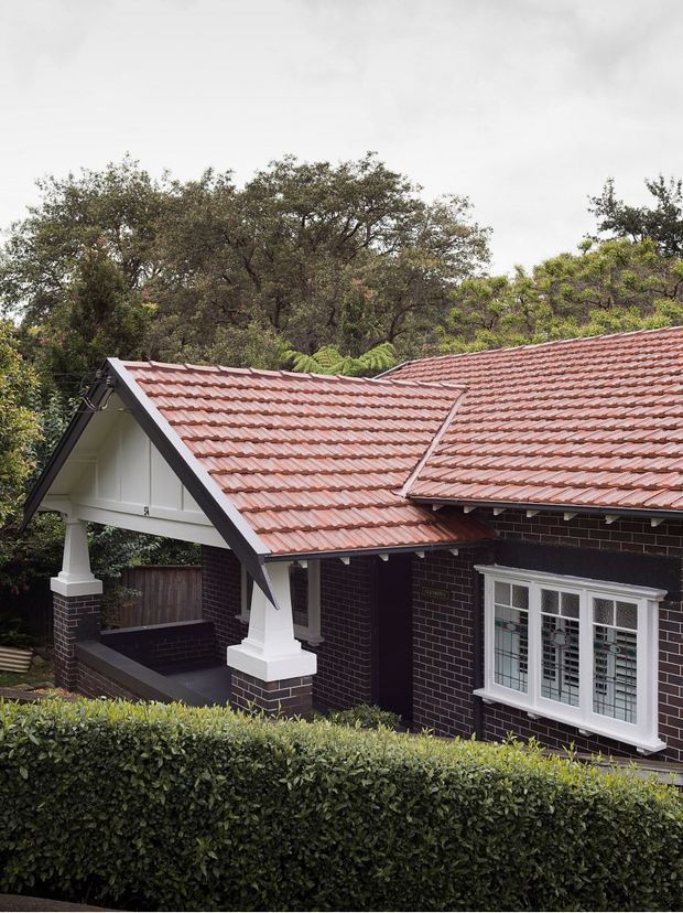 Exterior view of a traditional single-storey brick house, dominated by intersecting pitched roofs clad in terracotta-red tiles. The facade features dark brick walls, white timber gable detailing, decorative square columns, and a white multi-pane window with leadlight-style glazing. A neatly clipped hedge partially screens the lower facade, with mature trees behind the property under an overcast sky.