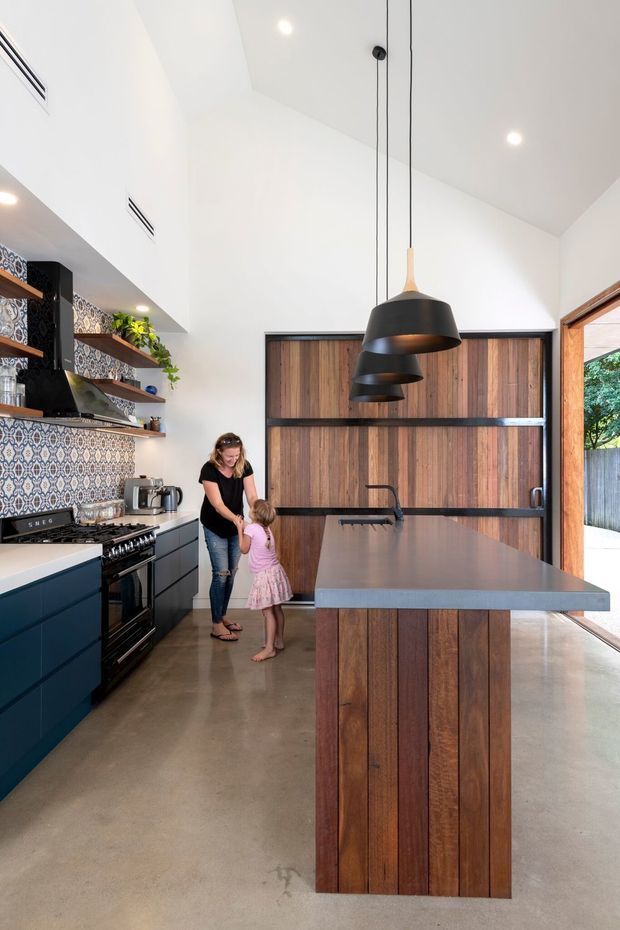 A bright, high-ceiling contemporary kitchen with a large central island, dark blue lower cabinetry, timber island cladding and timber-framed doors, patterned tile splashback, open timber shelving, and a row of black pendant lights. The kitchen opens to an outdoor area through large glazed doors. Polished concrete flooring and a dark stone or composite island benchtop complete the industrial-modern fit-out.