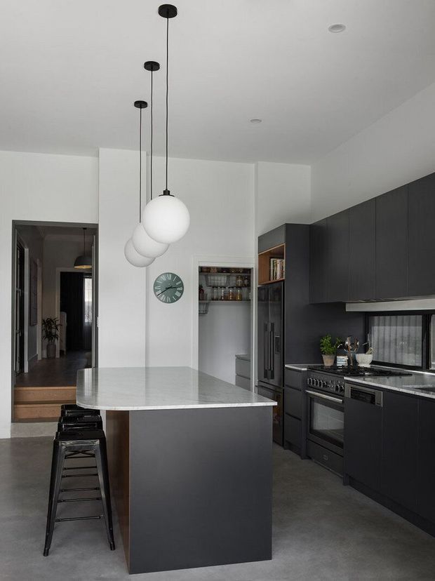 A contemporary kitchen with a dark charcoal island and full-height cabinetry, paired with a pale stone-look benchtop. Three white globe pendant lights hang above the island, while black stools provide seating. Integrated appliances, open shelving, a pantry nook and a small horizontal window complete the monochromatic scheme. A hallway is visible beyond the kitchen.