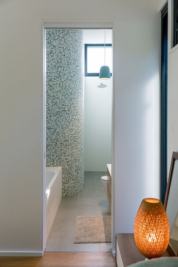 View through a doorway into a contemporary bathroom featuring a curved wall clad in small patterned mosaic tiles, a freestanding or built-in white bathtub, grey tiled flooring, a toilet, frosted glazing, and a teal pendant light. The foreground hallway includes a timber floor, white walls, dark-framed glazing, and a warm decorative lamp on a console.