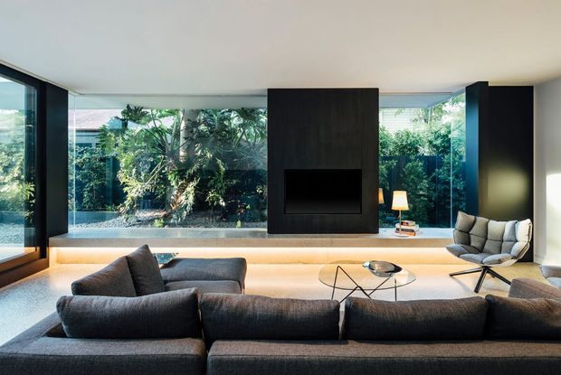 Contemporary living room with a large dark sectional sofa, sculptural glass coffee table, and freestanding lounge chair arranged around a central black fireplace feature wall. Full-height glazing opens the room visually to a lush courtyard garden, while warm concealed lighting runs beneath the fireplace and along the floor edge.