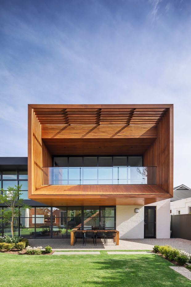 Contemporary two-storey residential facade viewed from the landscaped front yard. The upper level is framed by a deep timber-lined architectural opening with a slatted timber pergola and glazed balcony, while the lower level combines dark-framed glazing, a white masonry wall and an outdoor dining setting. The house has a strong indoor–outdoor connection and clean geometric composition.