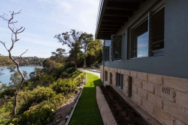 Exterior side view of a contemporary multi-level house on a landscaped waterfront or riverside site. The facade combines dark horizontal upper cladding and glazing with a textured pale stone masonry base. A narrow landscaped path and lawn run alongside the building, with water and vegetation visible beyond.