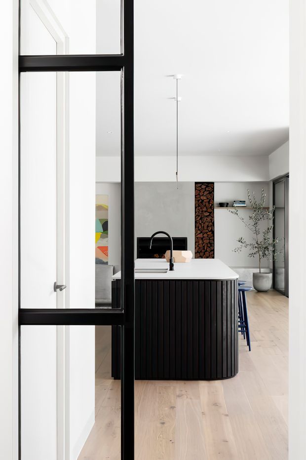 A bright contemporary kitchen viewed through a black-framed glazed door. The room features a white island with a fluted dark timber base, a black sink tap, pale timber flooring, white walls and cabinetry, a compact open wood-storage feature, pendant lighting, and a tall potted plant. Blue bar stools add a small accent of colour.