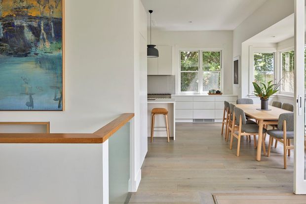 Bright open-plan interior showing a contemporary kitchen and dining area beyond a stair landing. The kitchen has white cabinetry, a pale tiled splashback, timber-look flooring, and a compact island with a timber stool. The adjacent dining space features a light timber table, upholstered chairs, and a potted plant. A timber handrail, abstract artwork, and white walls frame the foreground.