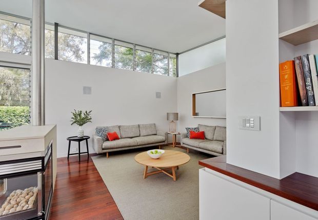 Bright contemporary living room with double-height proportions, extensive clerestory and full-height glazing, warm timber flooring, neutral upholstered sofas, a round timber coffee table, and built-in white joinery with open shelving and dark timber benchtops. Garden and trees are visible through the windows.