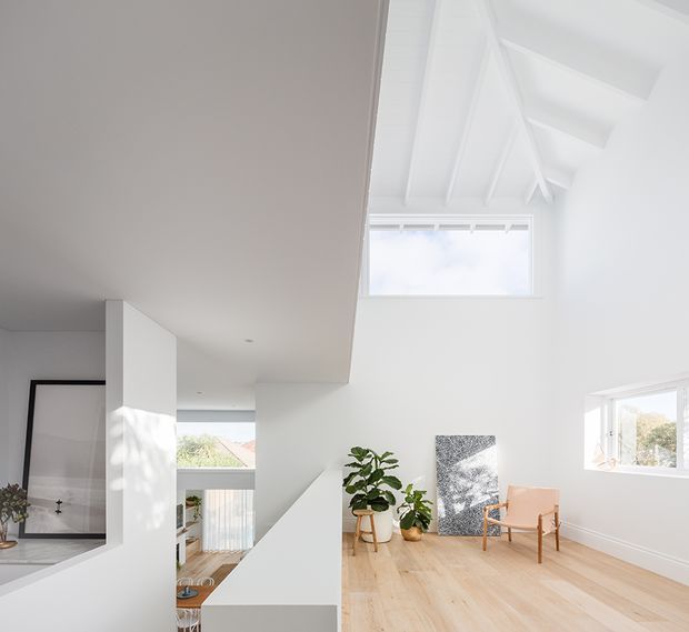 Bright, minimalist open-plan interior with white walls and a dramatic vaulted ceiling featuring exposed painted rafters. Pale timber flooring, abundant natural light from clerestory and side windows, a sculptural white stair or mezzanine structure, potted plants, a framed artwork, and a simple timber lounge chair create an airy contemporary living space.